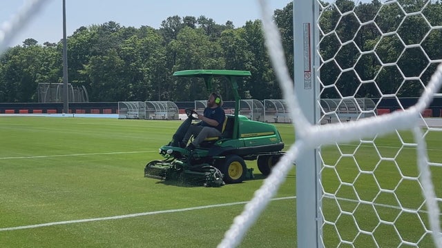 A man riding a lawn mower cuts the grass at a field at Stockton University, he's seen here through a bit of the netting of the goal 